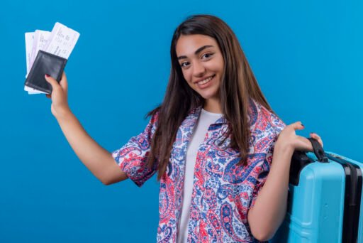 beautiful tourist woman holding travel suitcase and passport with tickets looking at camera with smile on face happy and positive, travel concept standing over blue background